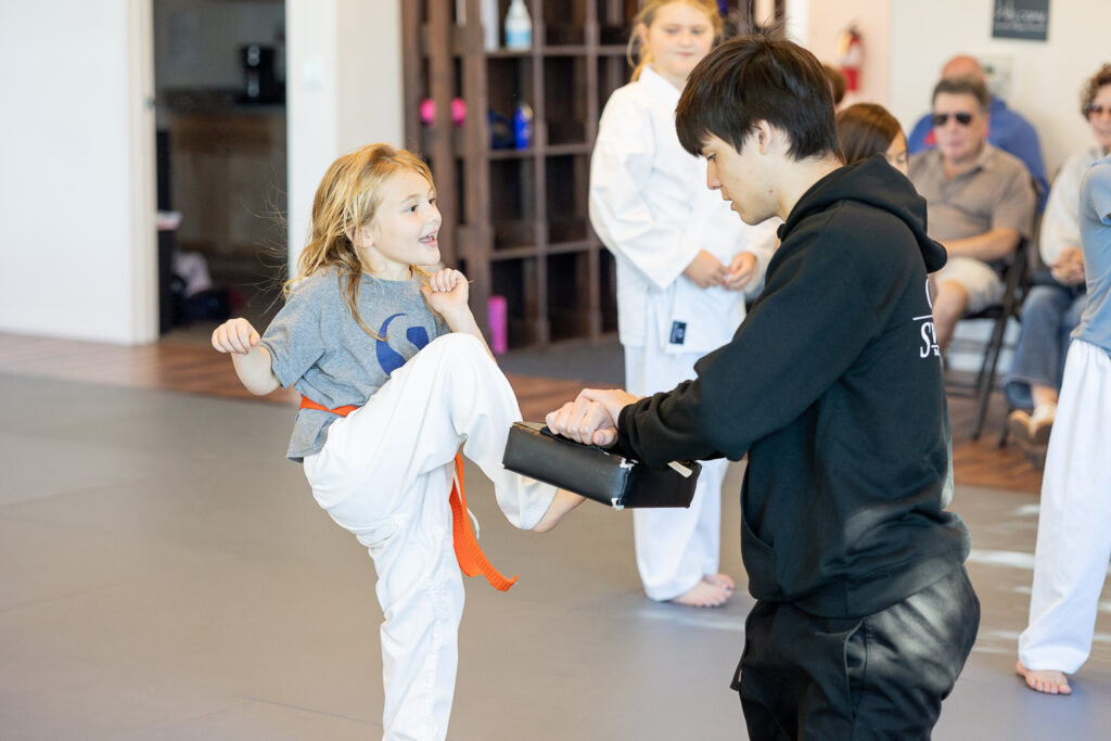 Little orange belt kicking a pad while an instructor assists her